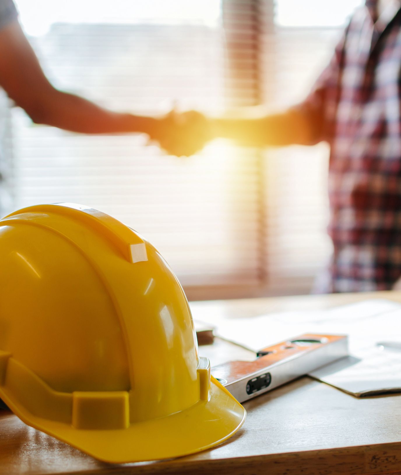 A yellow construction hard hat sits on a table with papers and tools, while two people in the background shake hands in a brightly lit room.