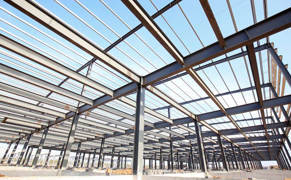 Steel framework of a large building under construction, with beams and columns forming a grid structure, open to the blue sky above.