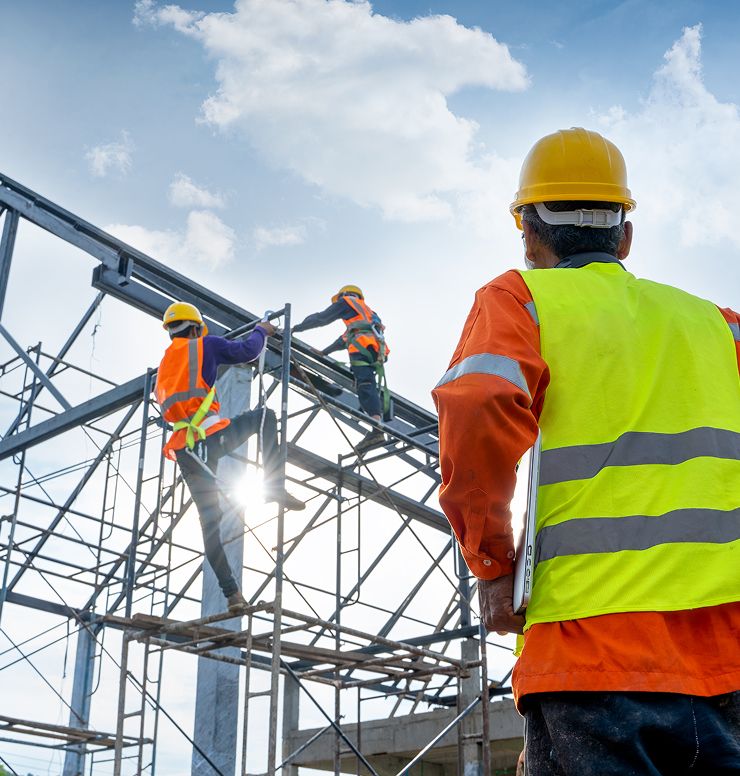 Construction workers wearing safety vests and helmets work on scaffolding and a steel structure under a blue sky, while another worker stands nearby observing the site.