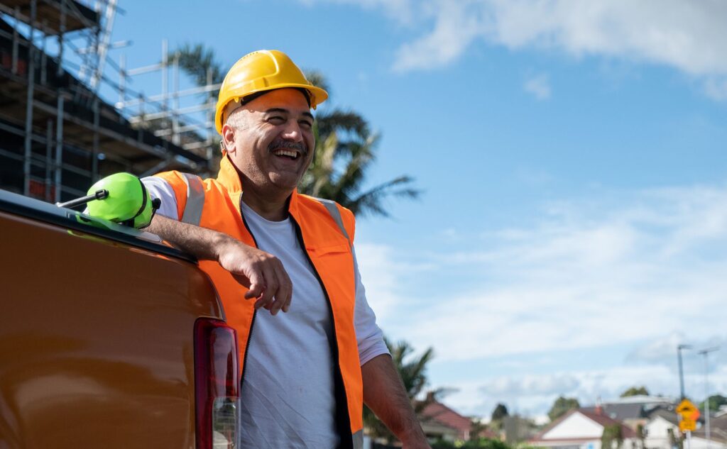 A smiling construction worker in an orange vest and yellow hard hat leans on the back of a truck at a building site, with scaffolding, palm trees, and houses visible in the background under a blue sky.