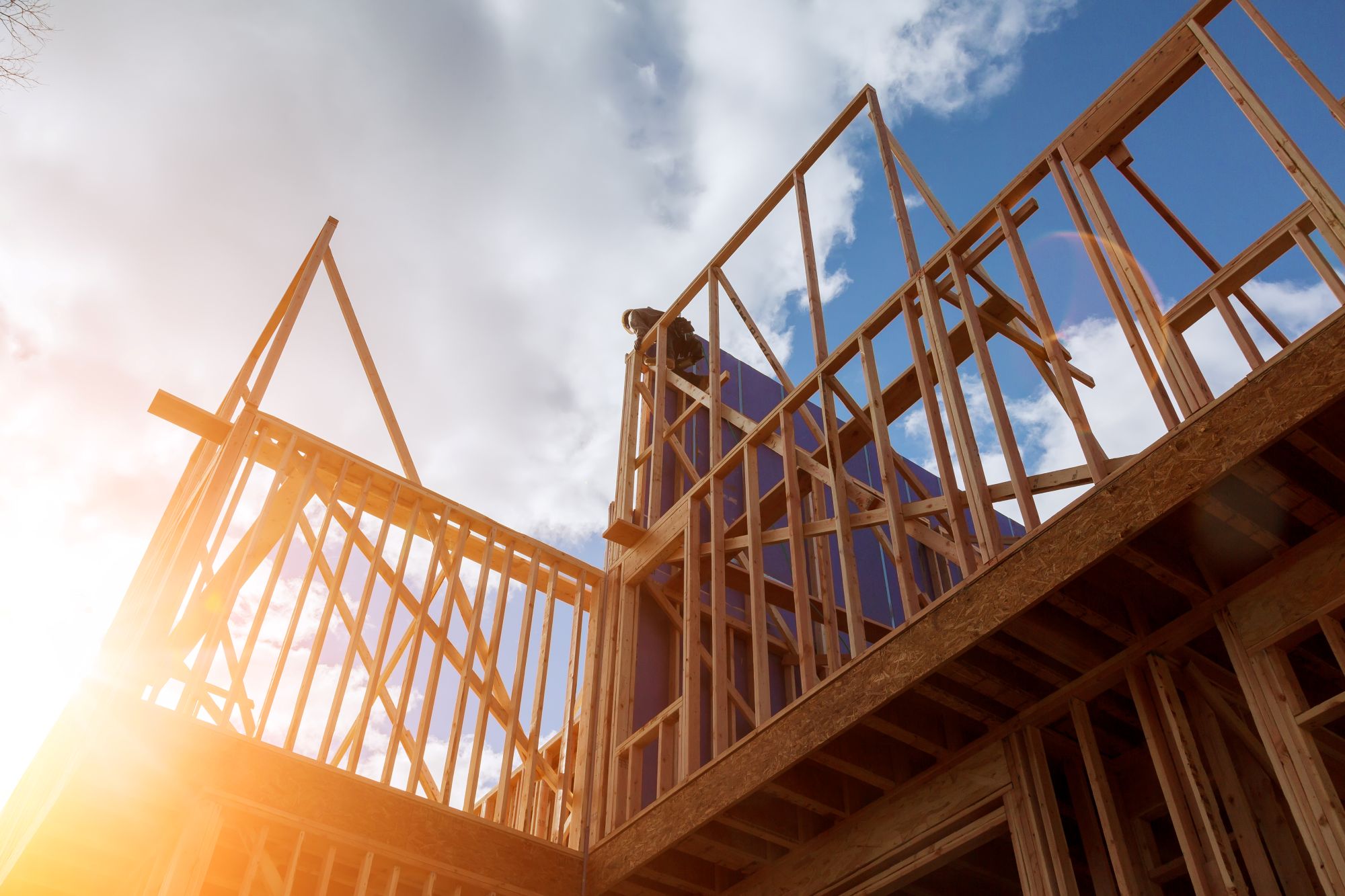 A low-angle view of a wooden house frame under construction, with sunlight streaming in from the left and partly cloudy sky above.