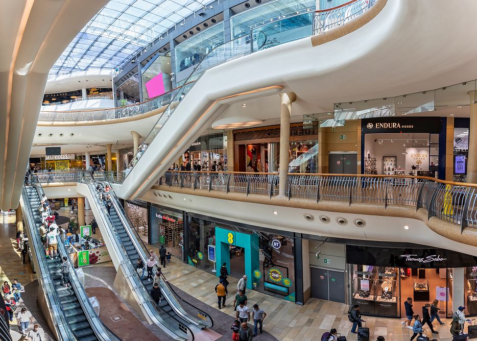 A busy, modern shopping mall with multiple floors, escalators, glass ceiling, and various stores. Shoppers are walking, browsing shops, and riding escalators in a bright, spacious environment.