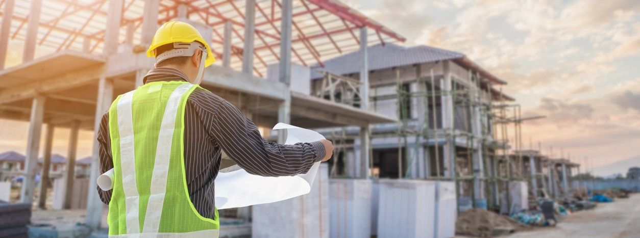 A construction worker wearing a yellow hard hat and reflective vest reviews blueprints at an active building site, with a partially constructed structure and scaffolding in the background.