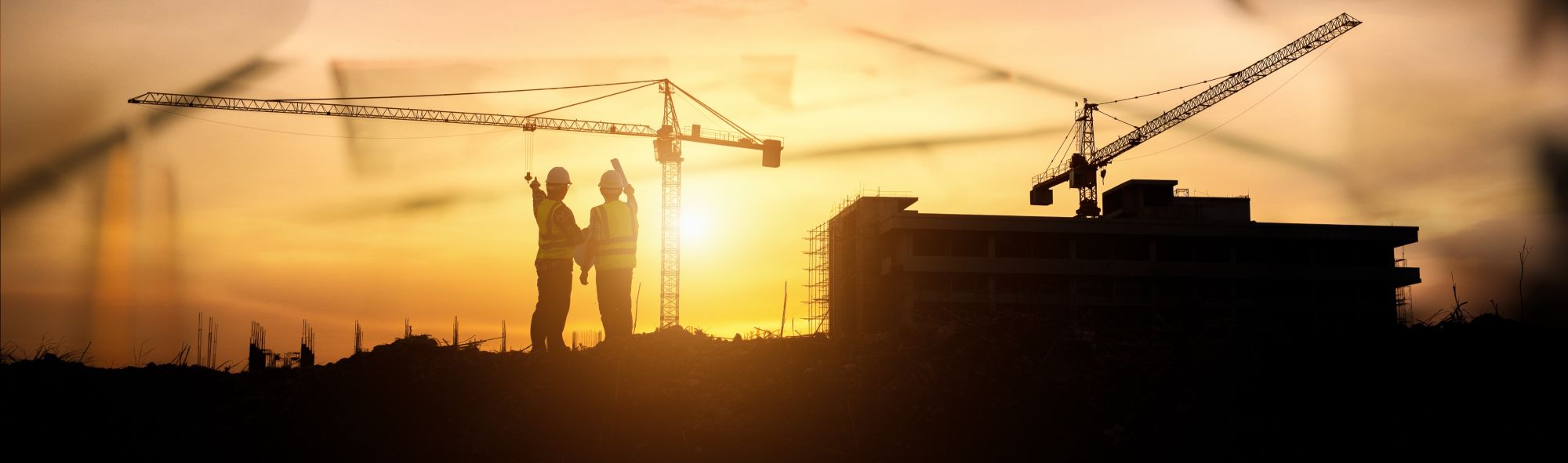 Silhouettes of two construction workers wearing safety vests and helmets stand near a building under construction with cranes, set against a sunset sky.