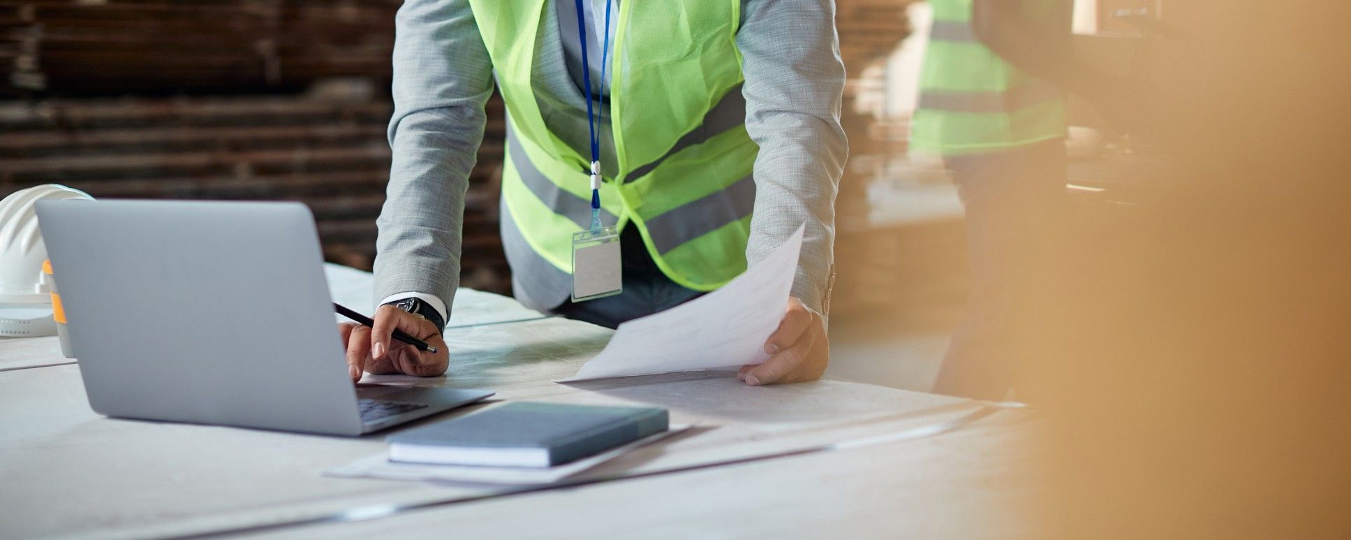 A person in a safety vest stands at a desk, using a laptop and holding documents, with a notebook nearby, suggesting a work or construction site setting.
